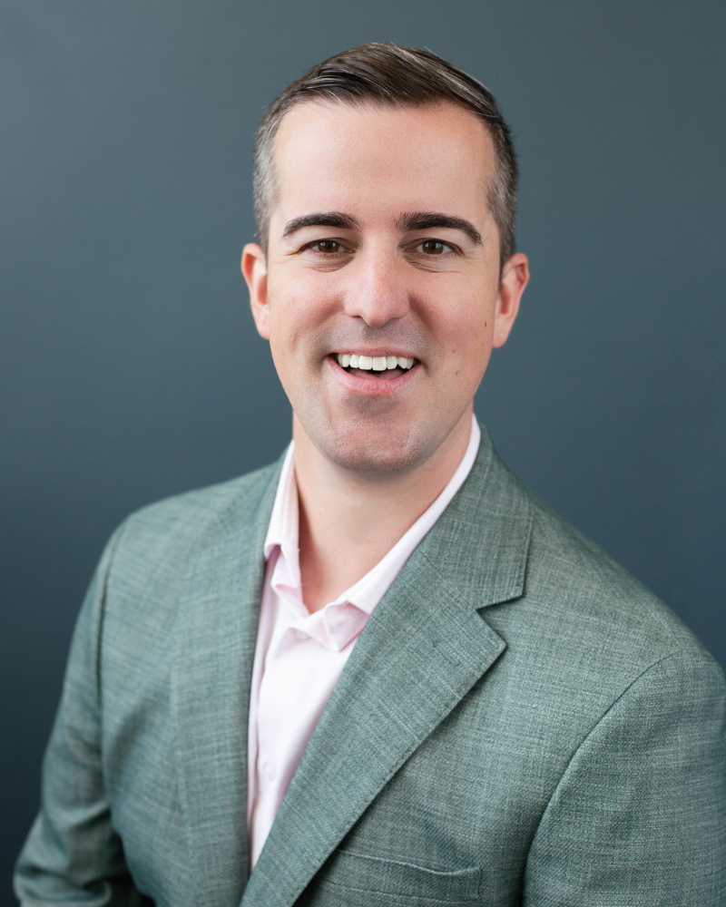 Professional headshot of a smiling man wearing a light green blazer and white shirt, photographed against a dark blue background.